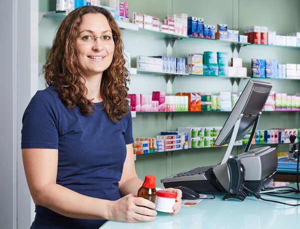 Moderne Innenansicht der Löwen-Apotheke, Apothekerin steht am Thresen mit einem Fläschchen und einem Medikament in der Hand.