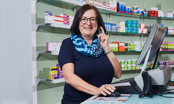 Apothekerin in einem blauen Shirt am PC, mit dem Telefon in der Hand, lächelnd vor einem Regal mit Medikamenten in der Löwen-Apotheke in Saarwellingen.