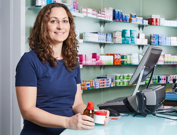 Moderne Innenansicht der Löwen-Apotheke, Apothekerin steht am Thresen mit einem Fläschchen und einem Medikament in der Hand.